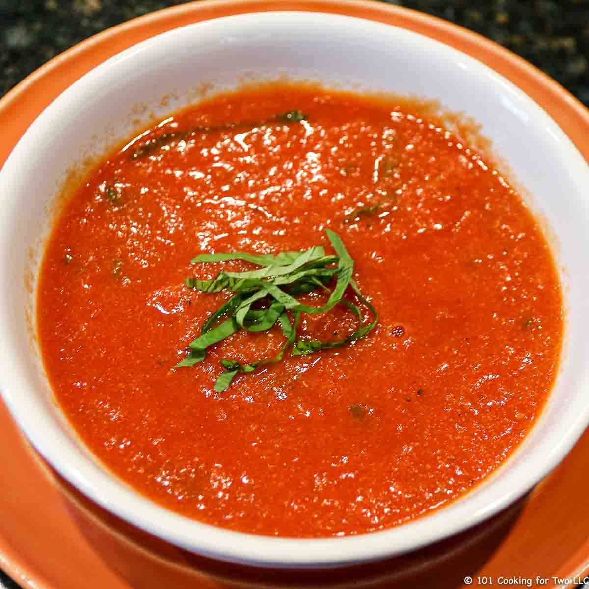 A bowl of tomato basil soup in a white bowl on orange plate.