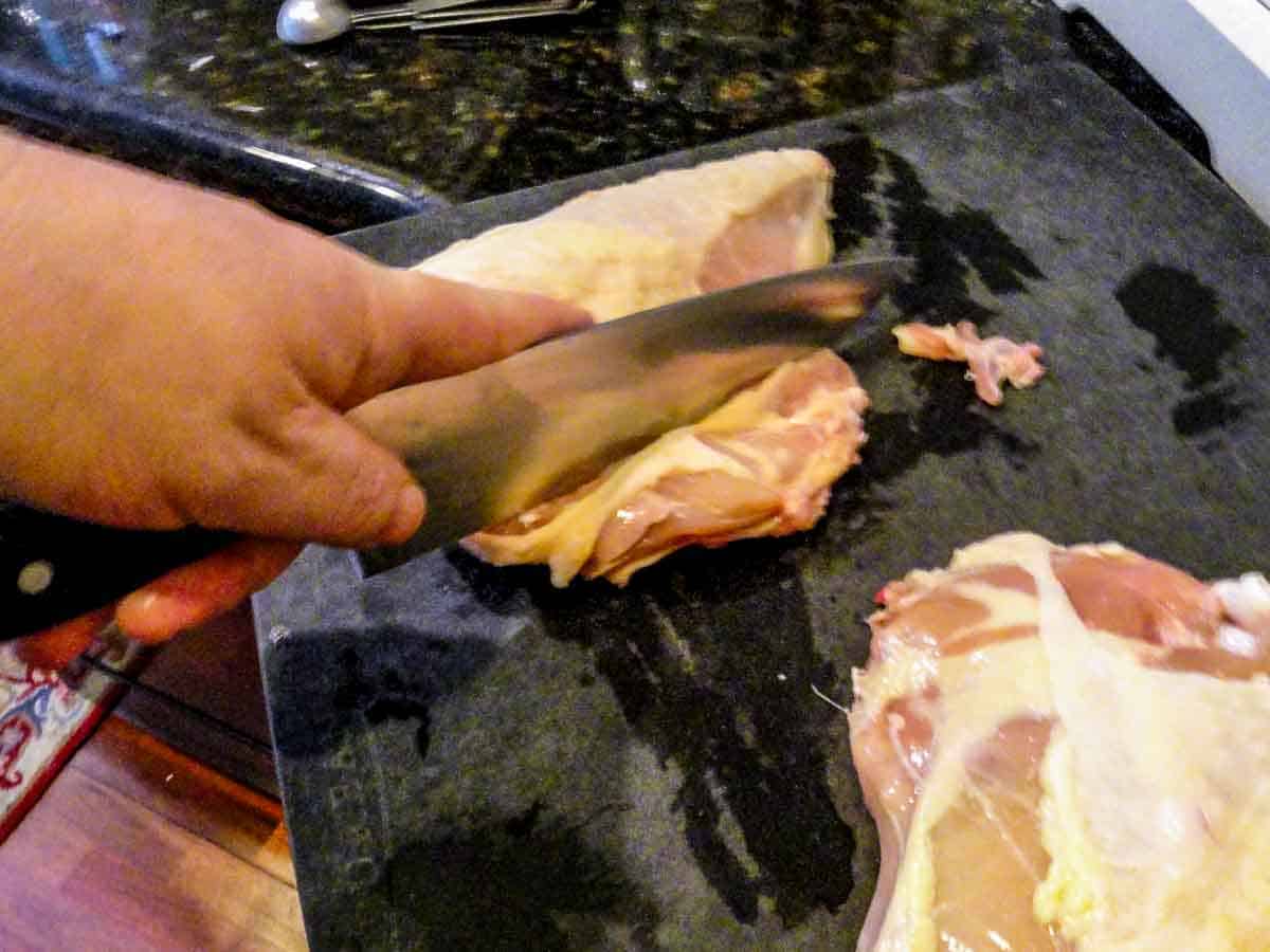 Cutting ribs off split chicken breasts with a chef knife on a black board