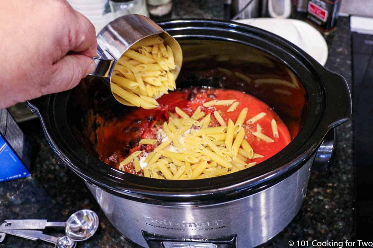 adding dry pasta to a crock pot with tomato sauce.