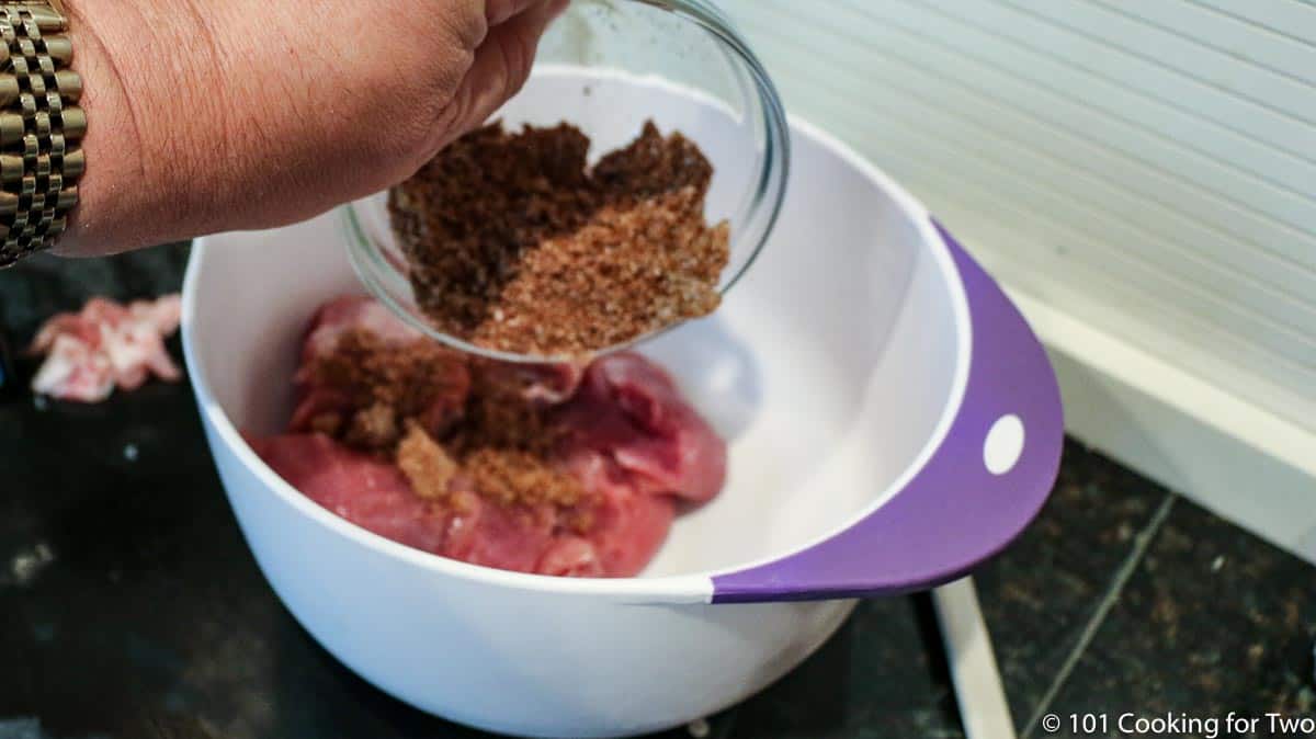 adding the mixed seasoning to a white bowl with chunks os pork tenderloin