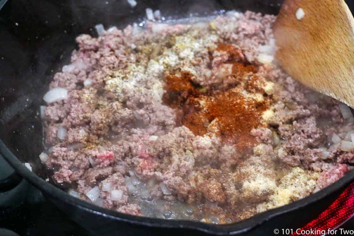 adding spices to raw burger while cooking in black pan