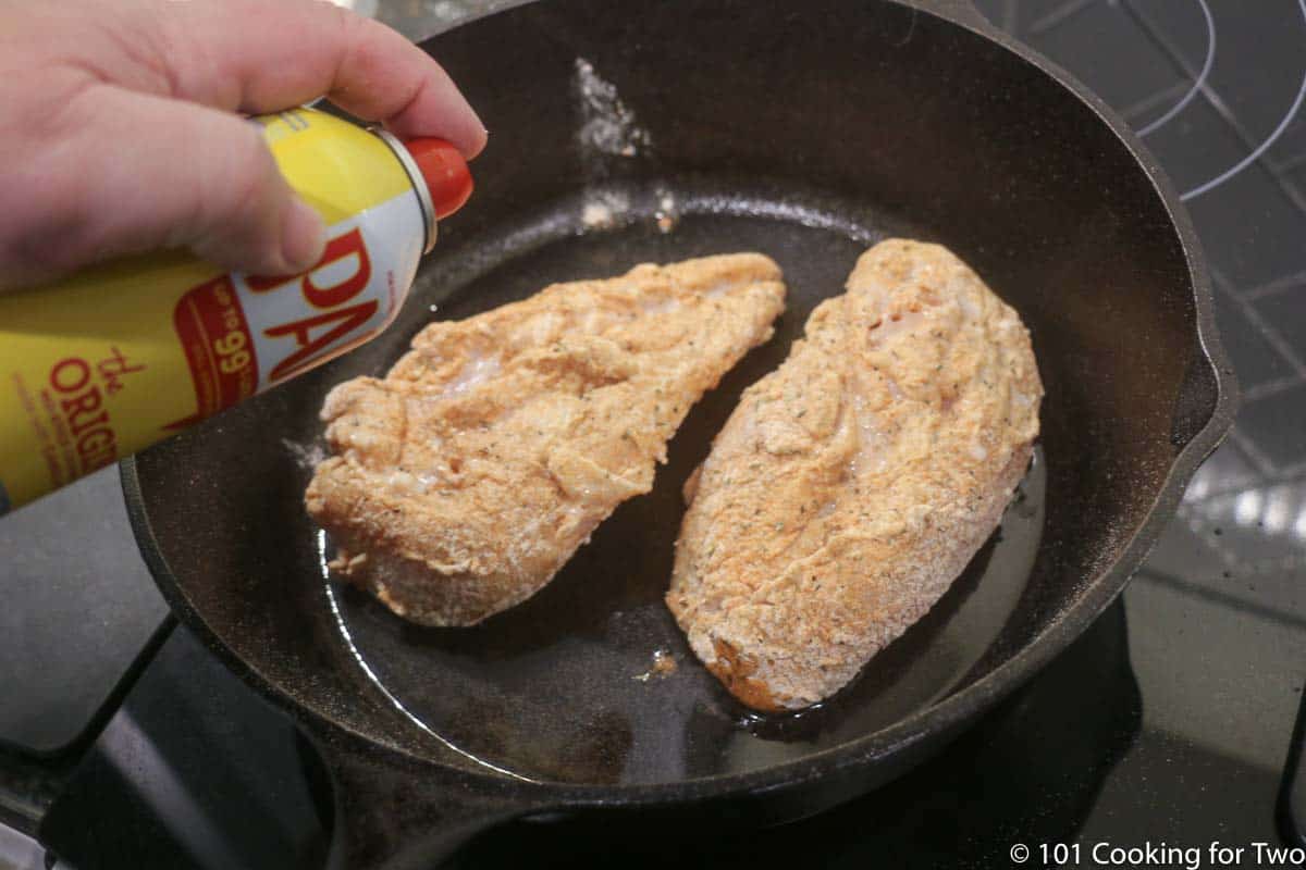 coated chicken breasts in pan being sprayed with PAM.