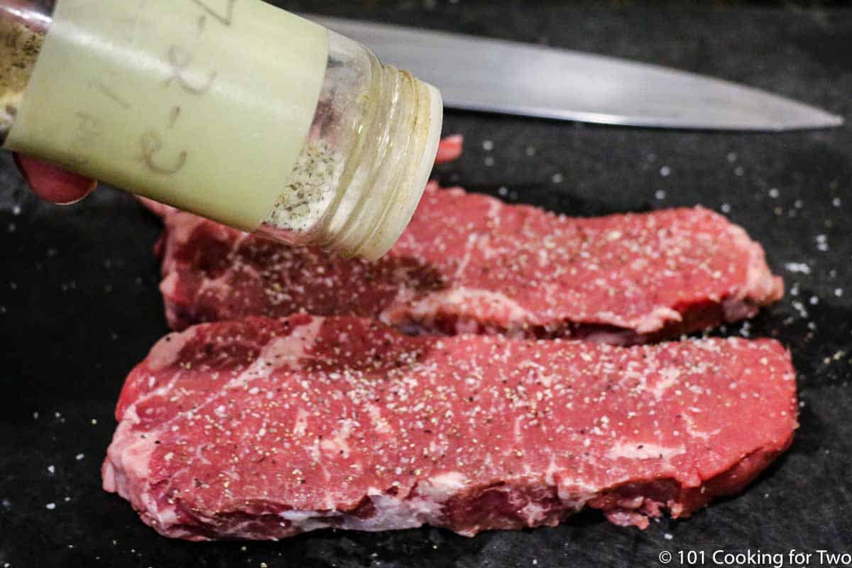 seasoning trimmed strip steaks on black board.