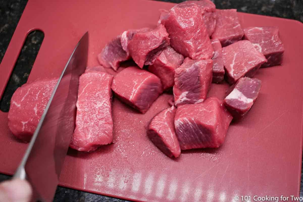 trimming steak into cubes on red board.