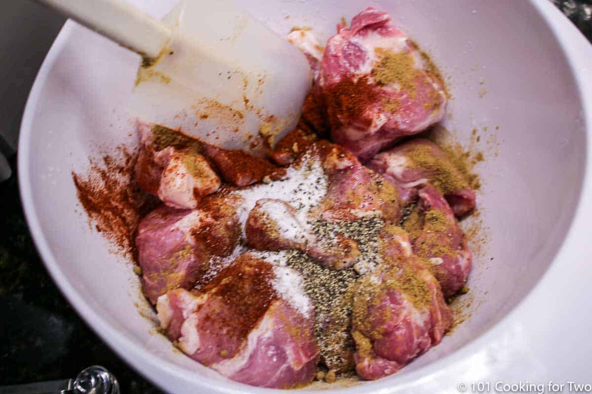 mixing chunks of pork butt with spices in a white bowl.