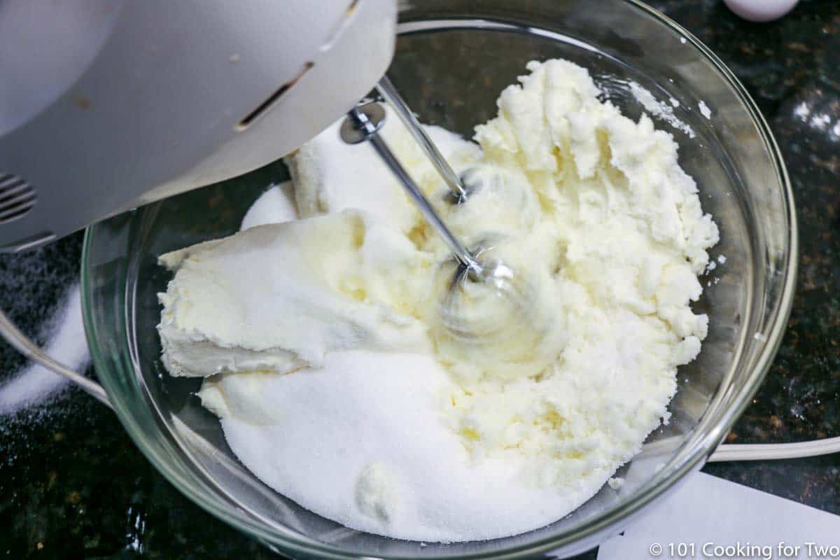 beating cream cheese with sugar in a glass bowl with an electric mixer