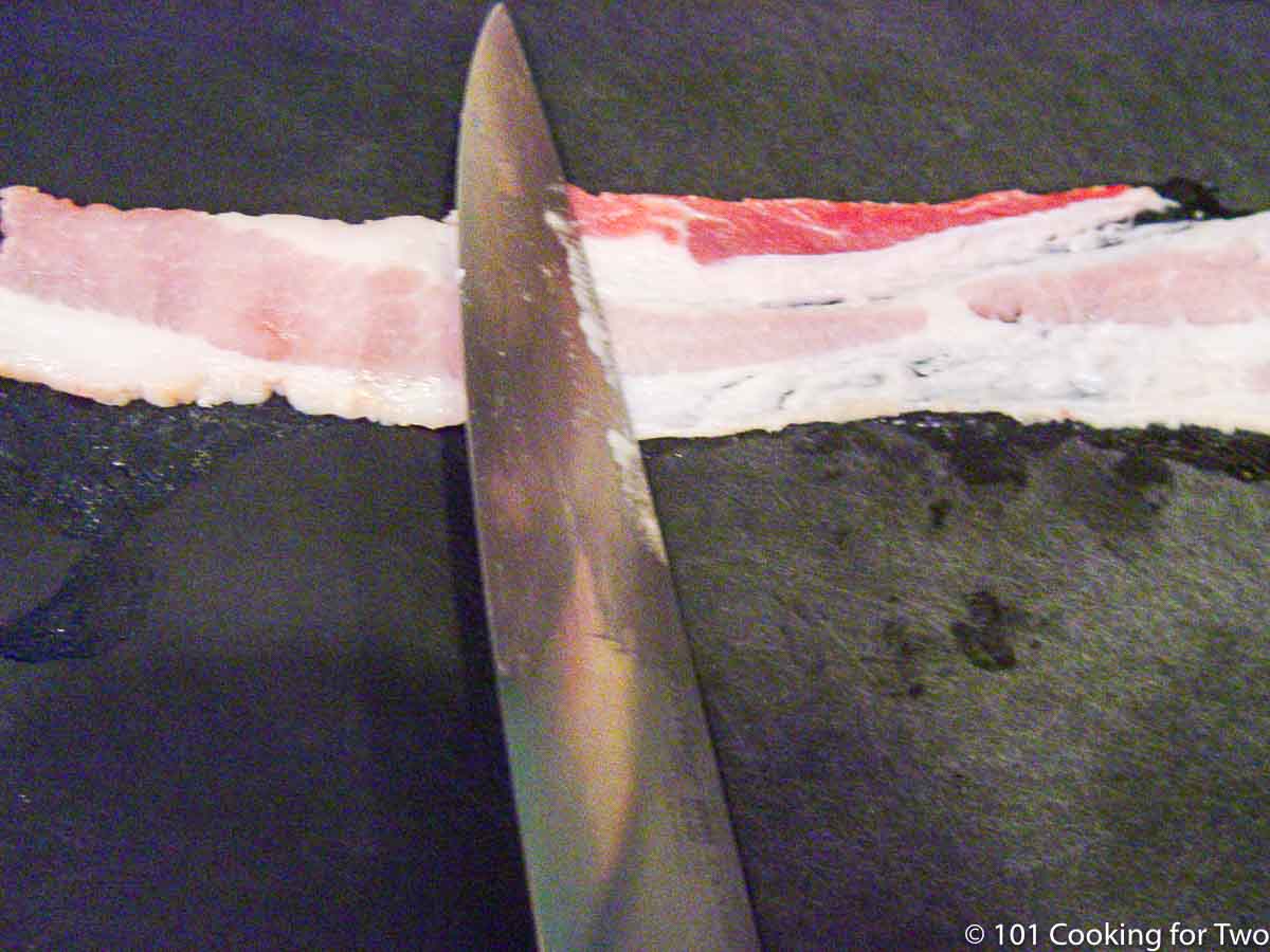flattening bacon strip on a black board with a knife.