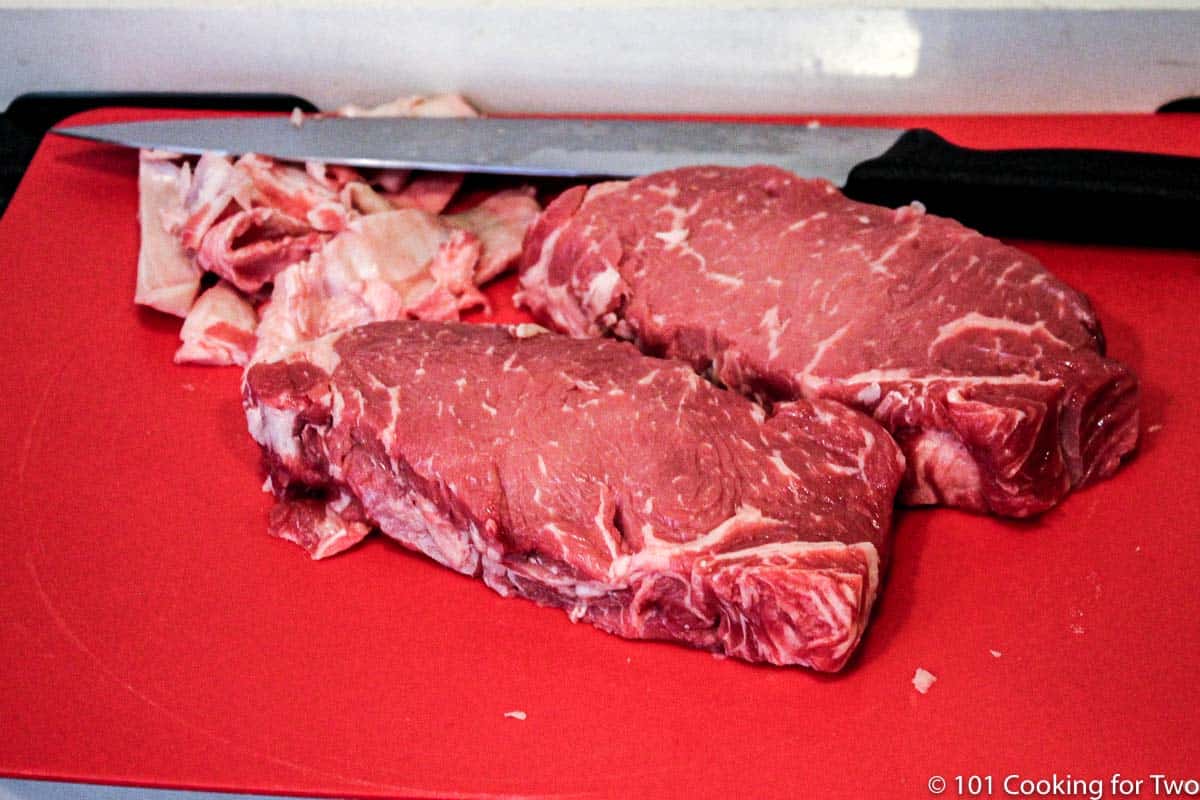 two trimmed New York Strip steaks on a red board.