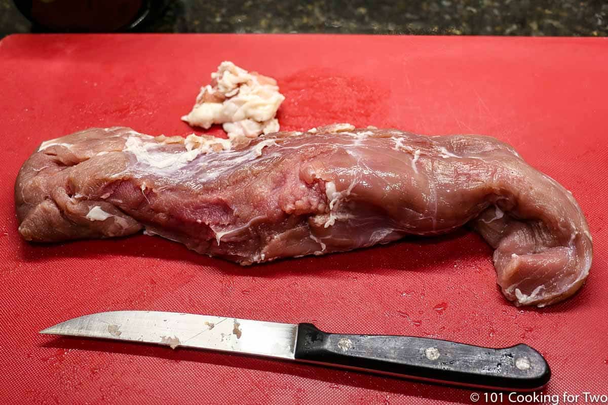 trimmed pork tenderloin on a red board with a knife.