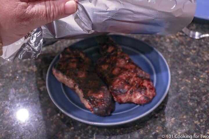 tenting strip steaks on a blue plate with foil