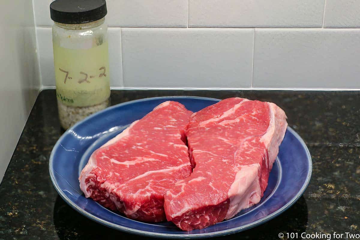 two NY strip steaks with seasoning on blue plate.