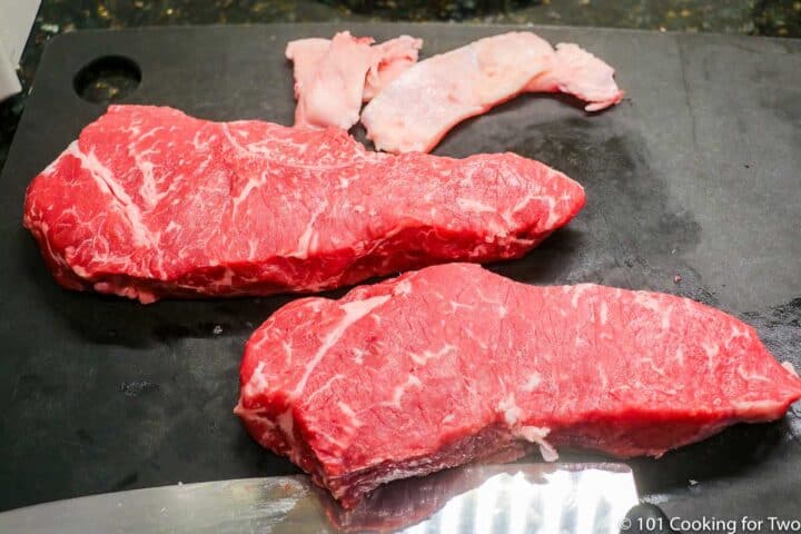 two trimmed strip steaks on a black board