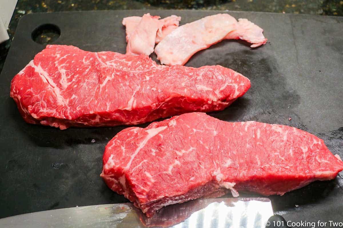 two trimmed strip steaks on a black board.