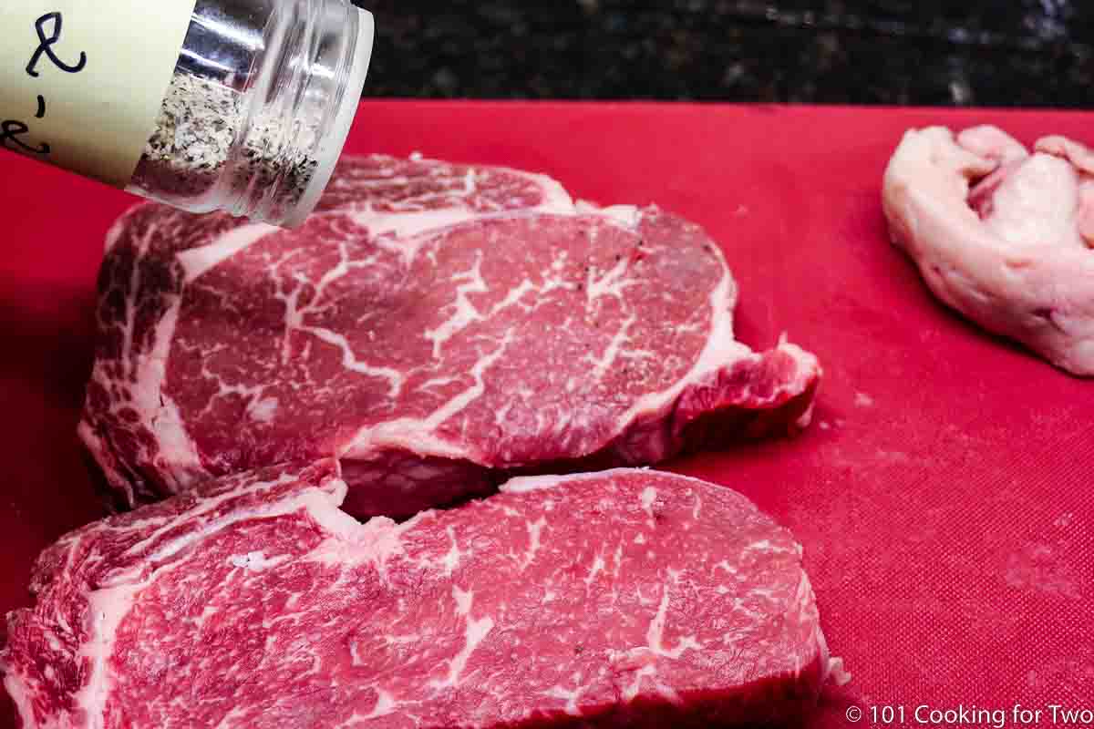 seasoning ribeye steaks on a red board.